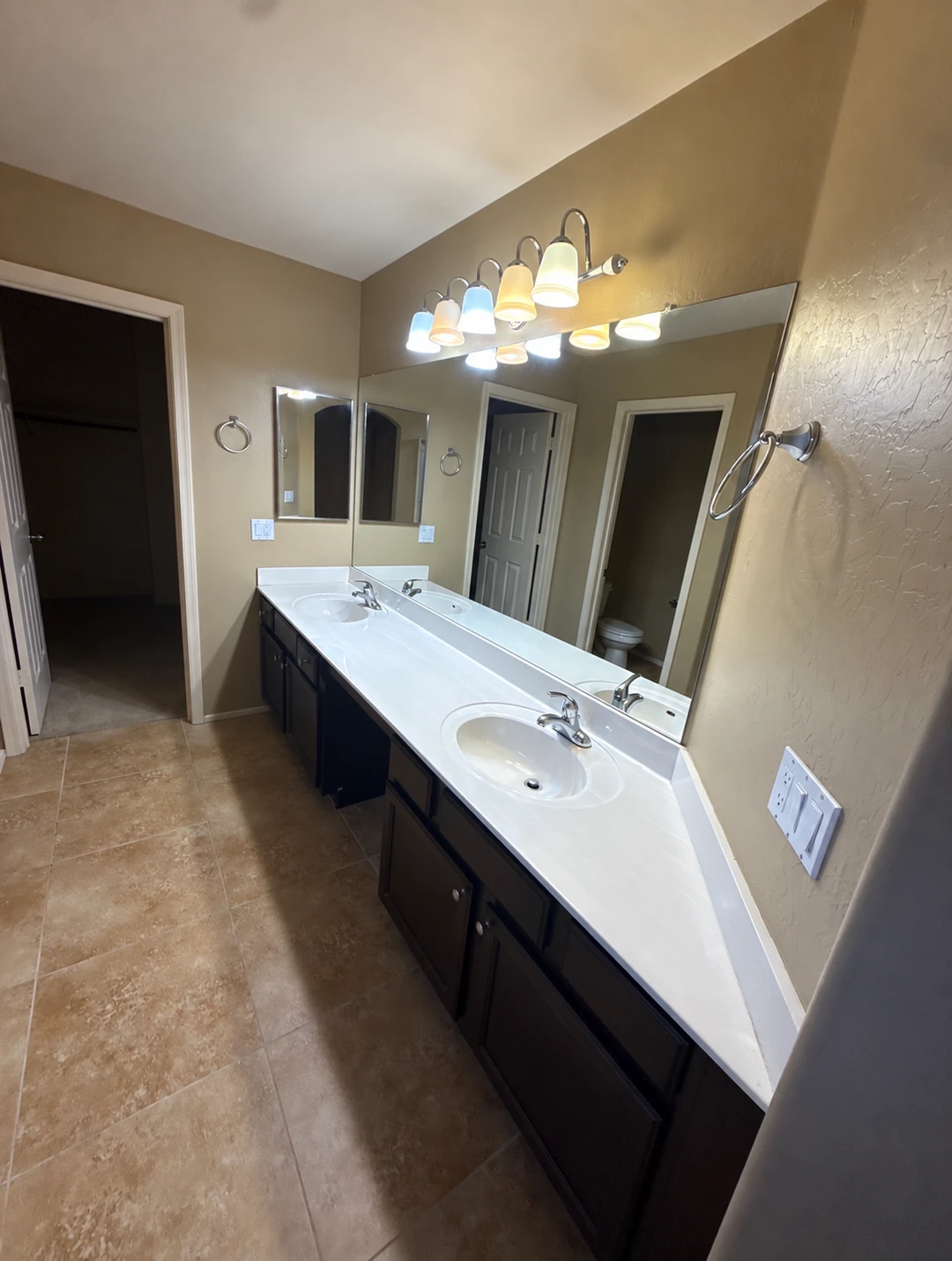 Bathroom before renovation, with a dark double vanity, white sinks, globe lights and beige tile